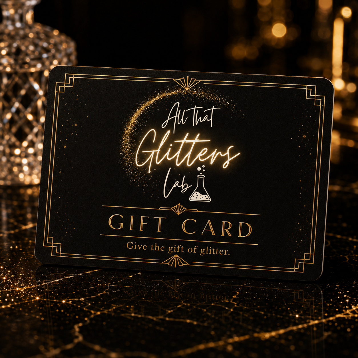 A black gift card with elegant gold glitter-style lettering and borders, reading 'All that Glitters Lab GIFT CARD' and 'Give the gift of glitter.' The card is photographed on a dark reflective surface with warm bokeh lights in the background.